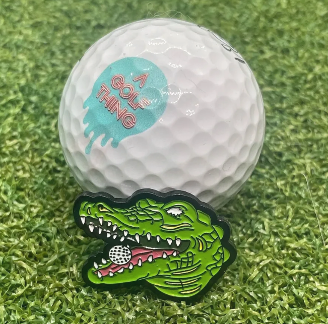 En vit golfboll med texten 'A GOLF THING' och en färgstark markör i form av en grinande krokodil framför den, på en konstgjord grön gräsmatta.