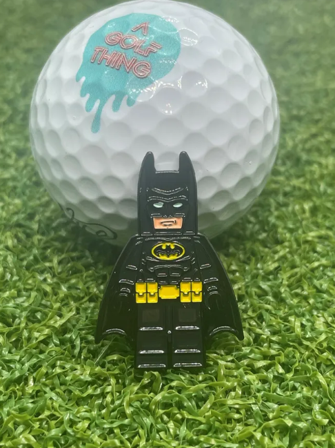 En golfbollmarkör av Batman i svart kostym står framför en vit golfboll med texten 'A GOLF THING' på en konstgjord grön gräsmatta.