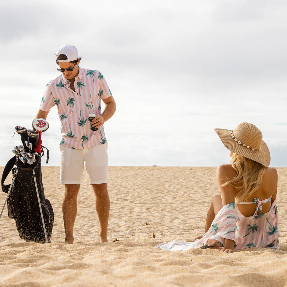 En golfare står med en golfbag på en vit sand, en kvinna sitter ner och kollar på golfaren. 